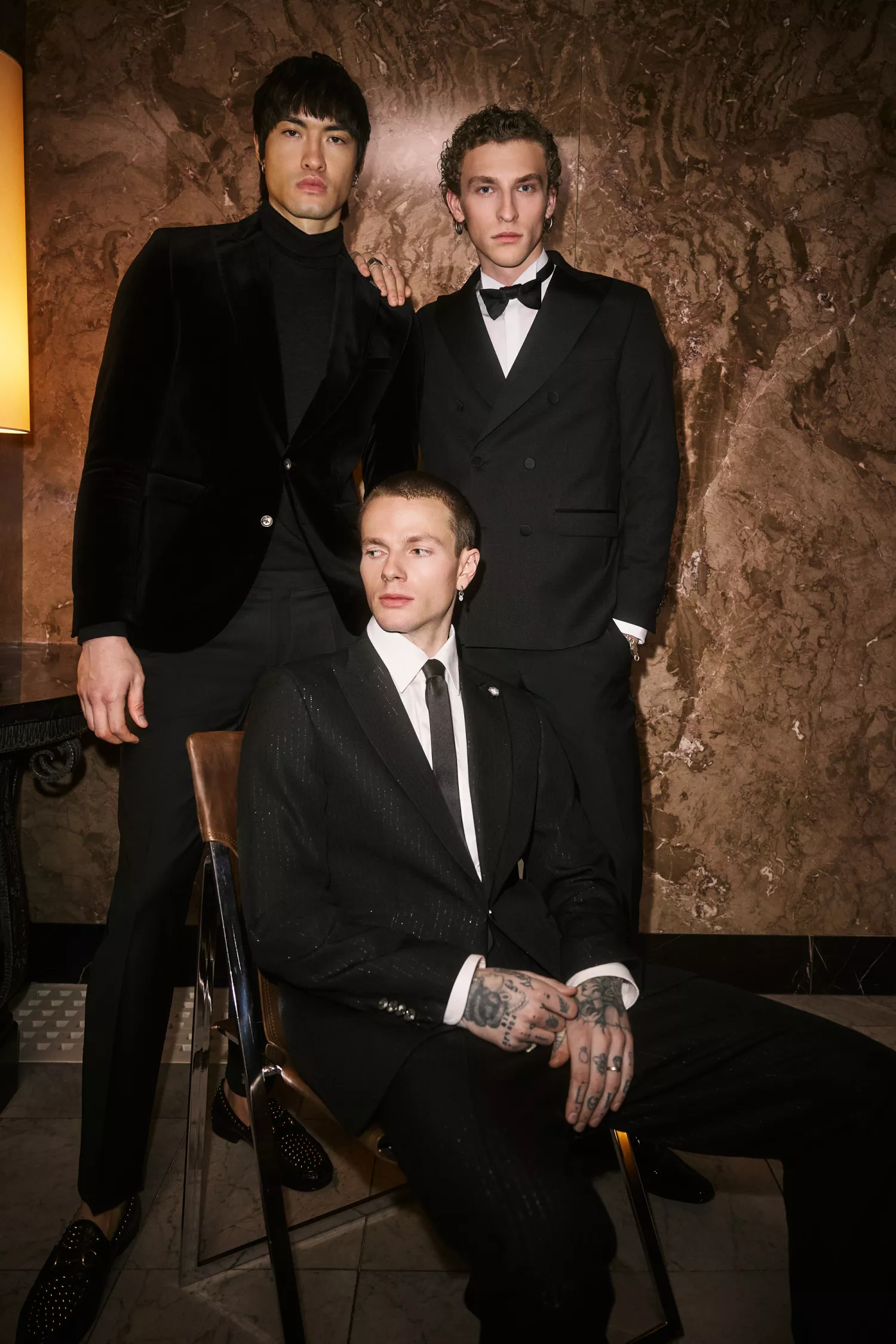Drei Modelle in eleganten schwarzen Anzügen und Smokings vor edler Marmorwand